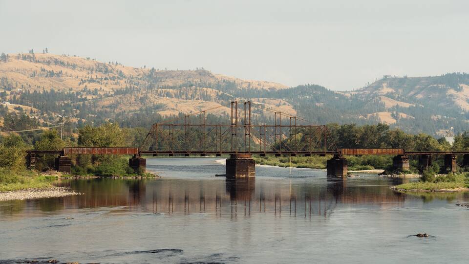 A river with a rusted bridge spanning across it, surrounded by hills and trees.