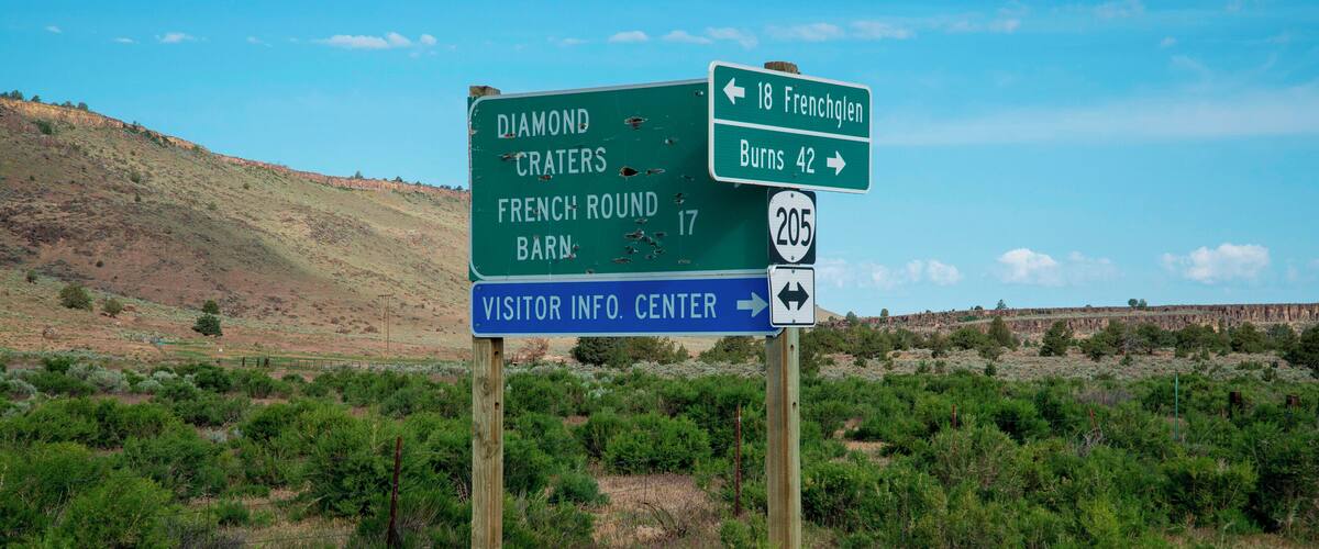 A directional sign in a desert landscape with green shrubs and a hill in the background.