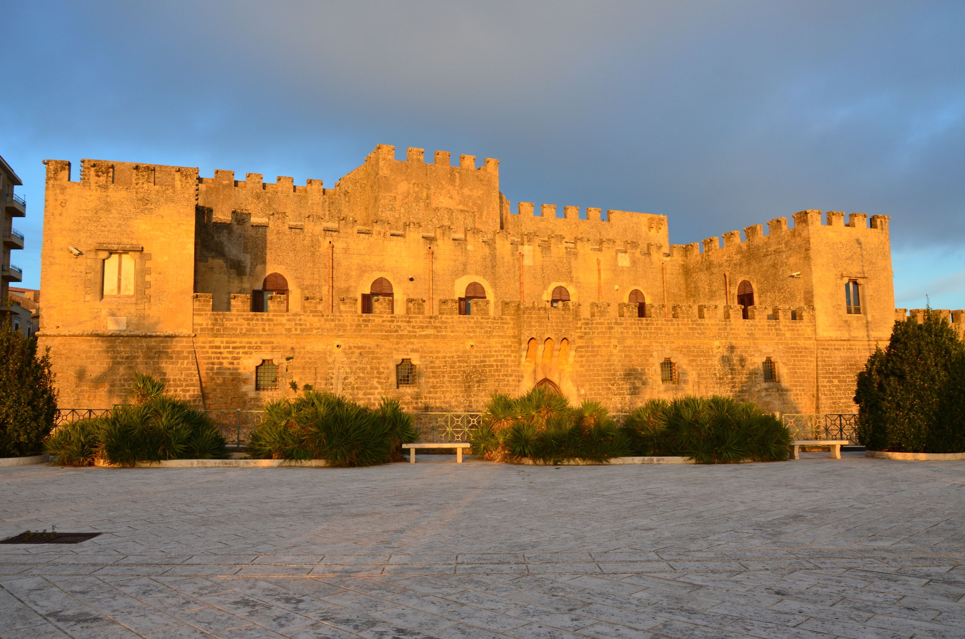 A historic stone castle with arched windows and battlements, surrounded by a paved courtyard and greenery.