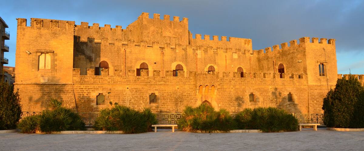 A historic stone castle with arched windows and battlements, surrounded by a paved courtyard and greenery.