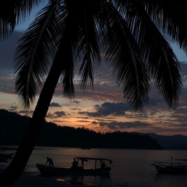 Um pôr do sol sobre um lago calmo, com barcos, palmeiras e uma linha de costa distante.
