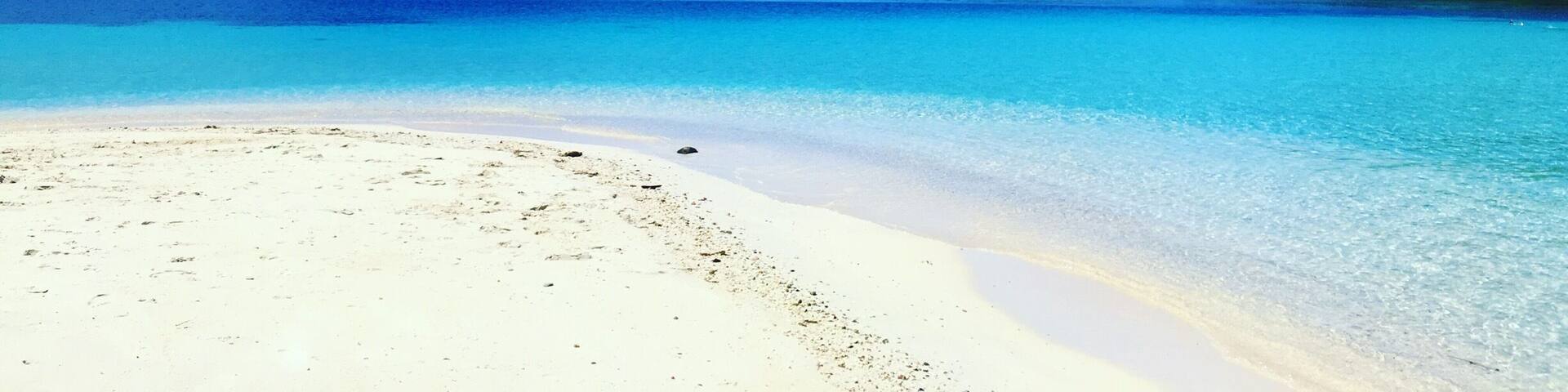 A beach with clear turquoise water, white sand, and green hills in the background.