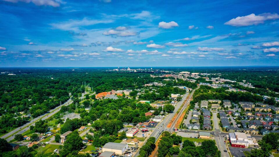 An aerial view of a town with a central road, surrounding greenery, and distant buildings.