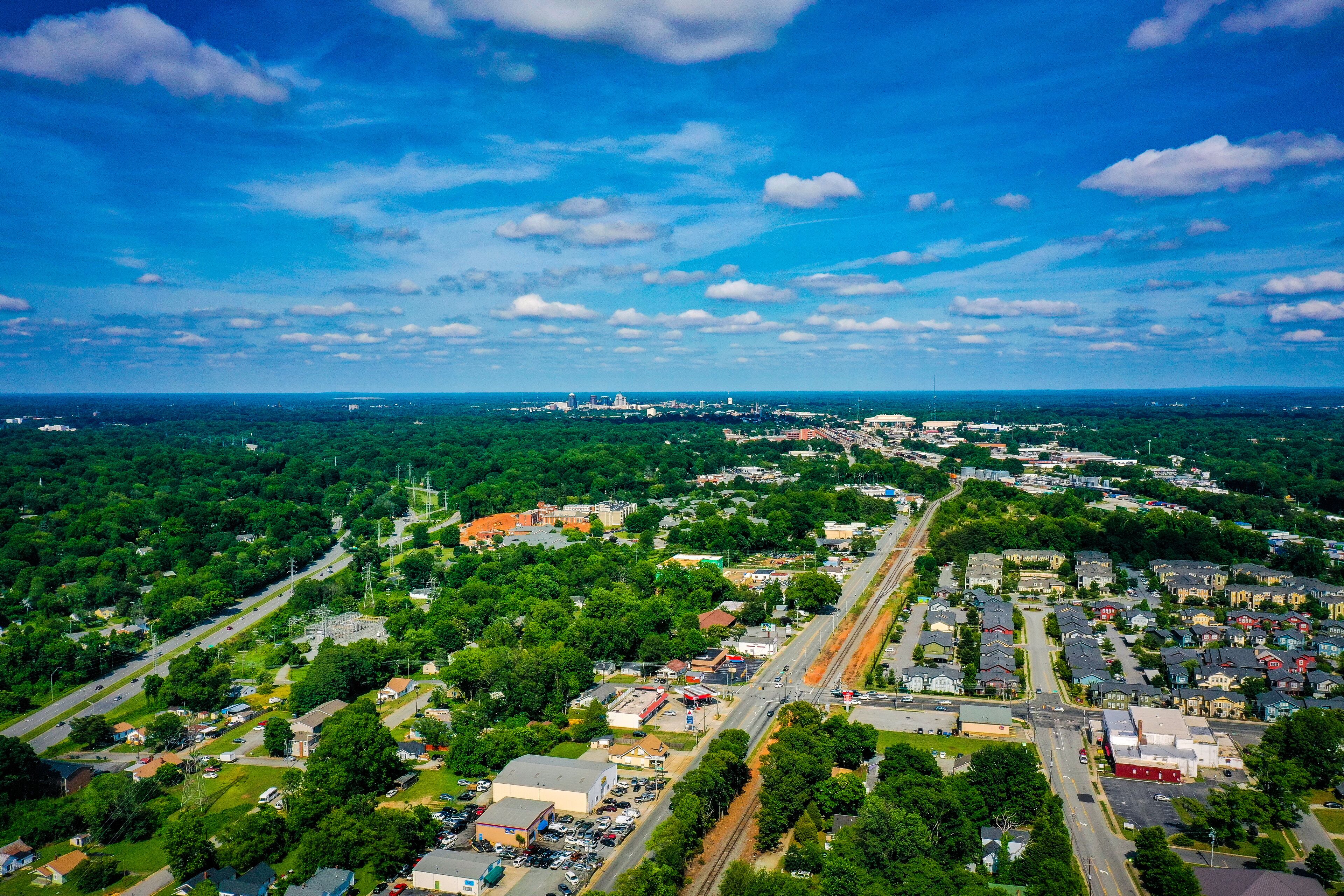 An aerial view of a town with a central road, surrounding greenery, and distant buildings.