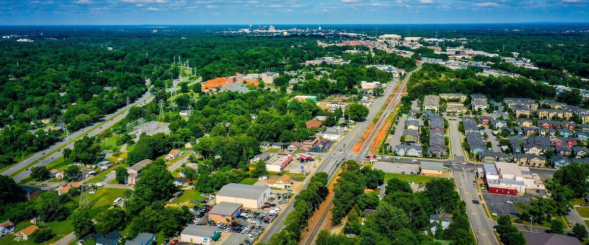 An aerial view of a town with a central road, surrounding greenery, and distant buildings.