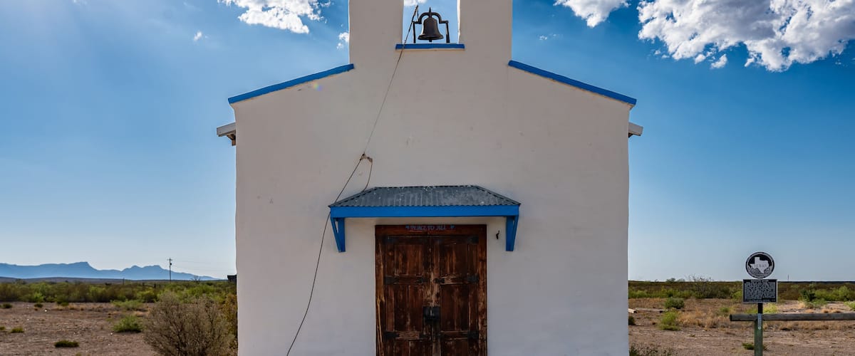 A white church with a blue roof and bell tower, set against a desert landscape with mountains in the background.