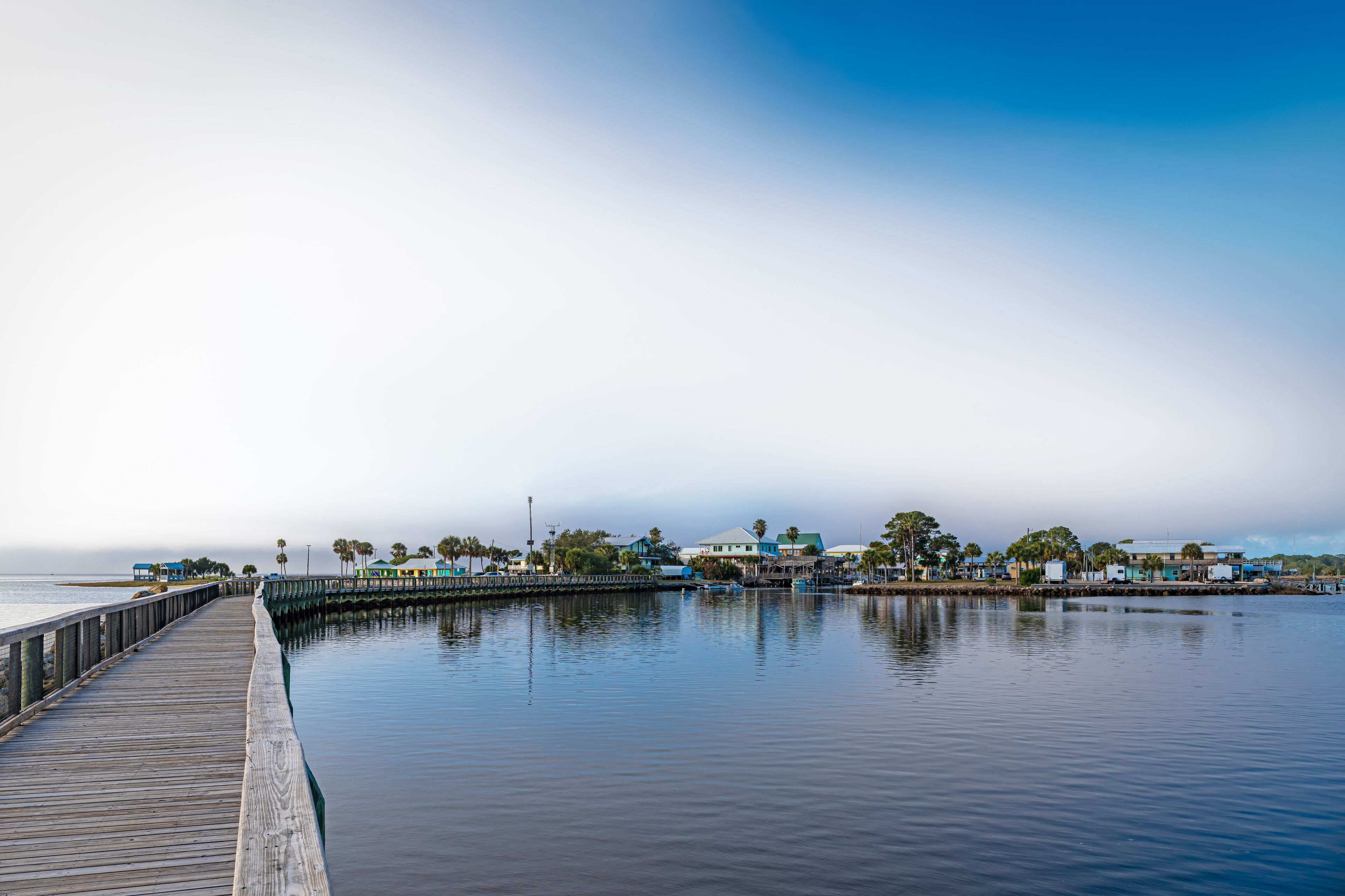 A wooden pier extending into calm water, with buildings and palm trees in the background under a clear sky.