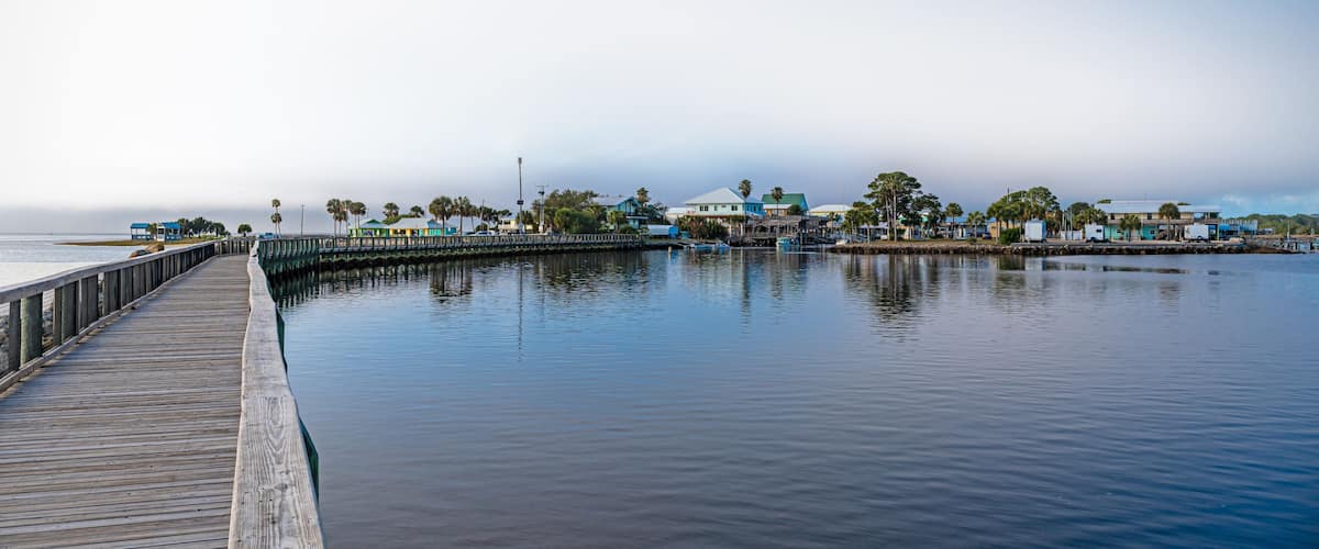 A wooden pier extending into calm water, with buildings and palm trees in the background under a clear sky.