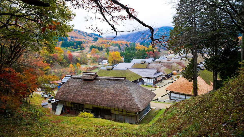 A village with traditional thatched-roof houses, surrounded by autumn-colored trees and hills in the background.