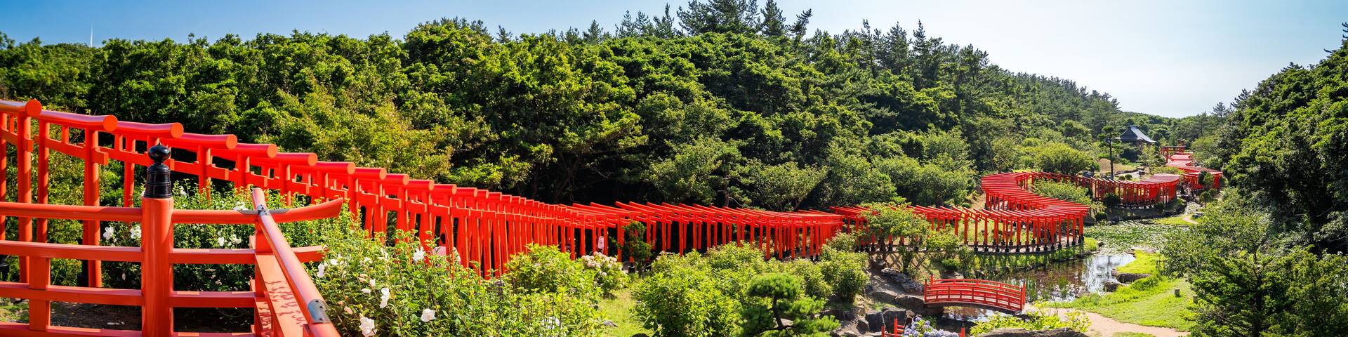 A red torii gate pathway leading through a lush green forest with a traditional Japanese building in the background.