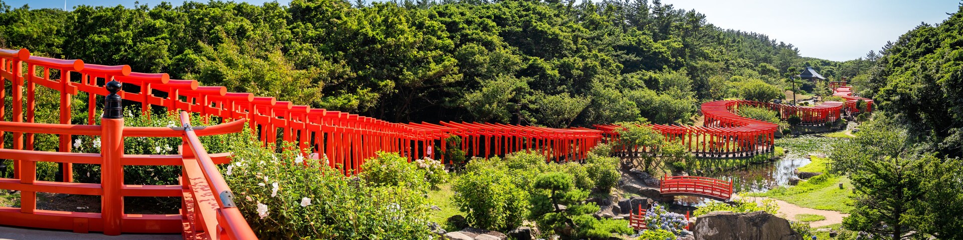 A red torii gate pathway leading through a lush green forest with a traditional Japanese building in the background.