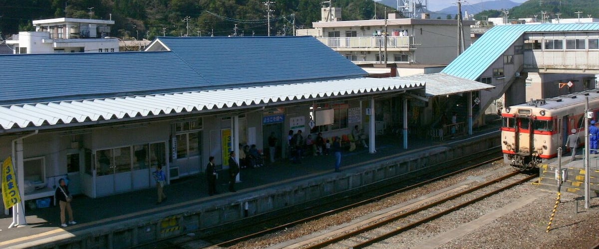A train station with a blue roof, passengers waiting, and a train approaching.
