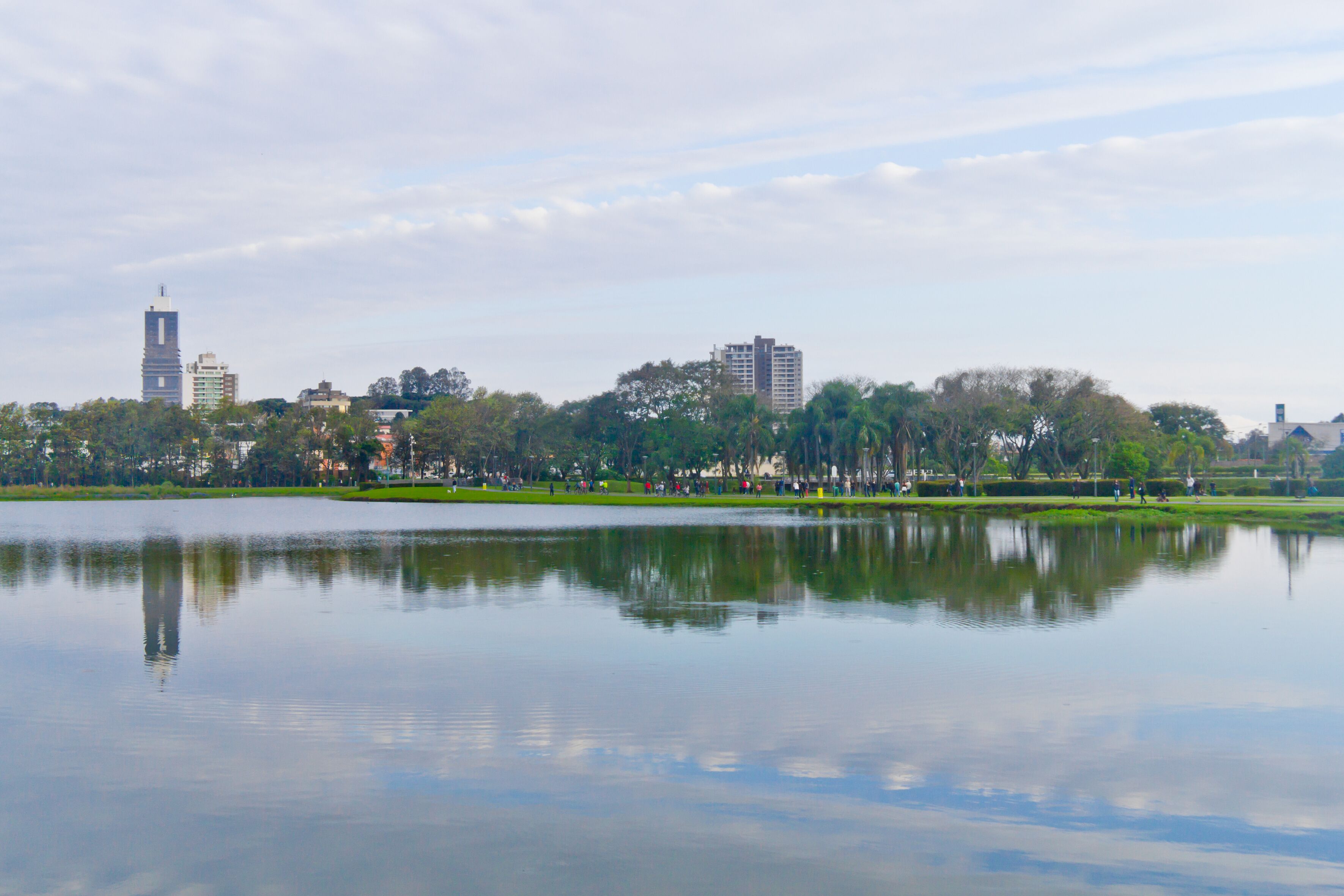 A calm lake reflecting buildings and trees, with a park in the foreground and a city skyline in the background.