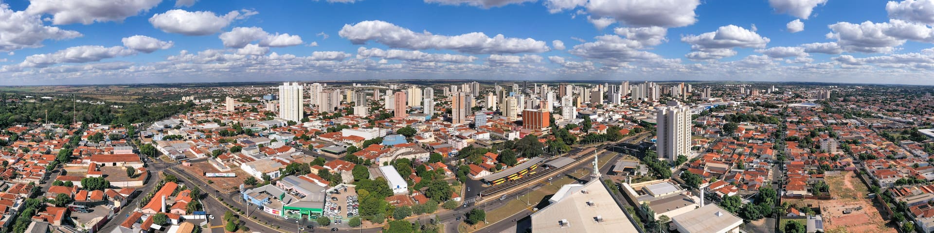 A cityscape with a mix of high-rise buildings and smaller houses, a clear blue sky with scattered clouds, and a distant view of a flat landscape.