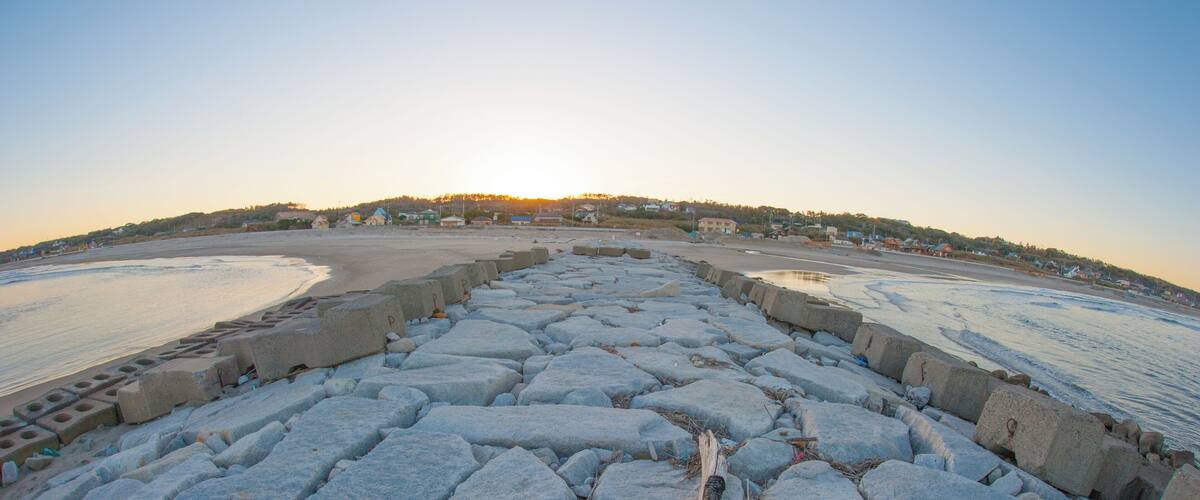 A rocky shoreline with a breakwater extending into the sea, a clear sky, and a distant village on a hill.