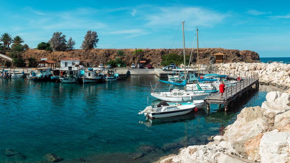 A coastal marina with boats docked, rocky shore, and buildings in the background.