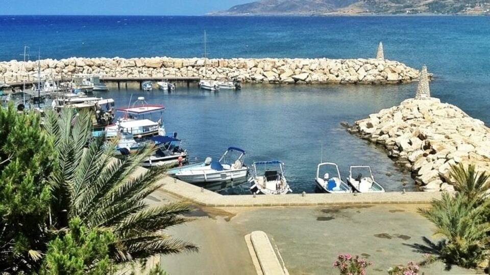 A marina with boats docked, a rocky breakwater, and a clear blue sea with distant hills.
