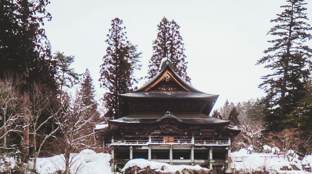 View of a traditional Japanese temple perched atop rocky cliffs, surrounded by snow-dusted trees and a serene, wintry landscape, Yanaizu, Fukushima, Japan.