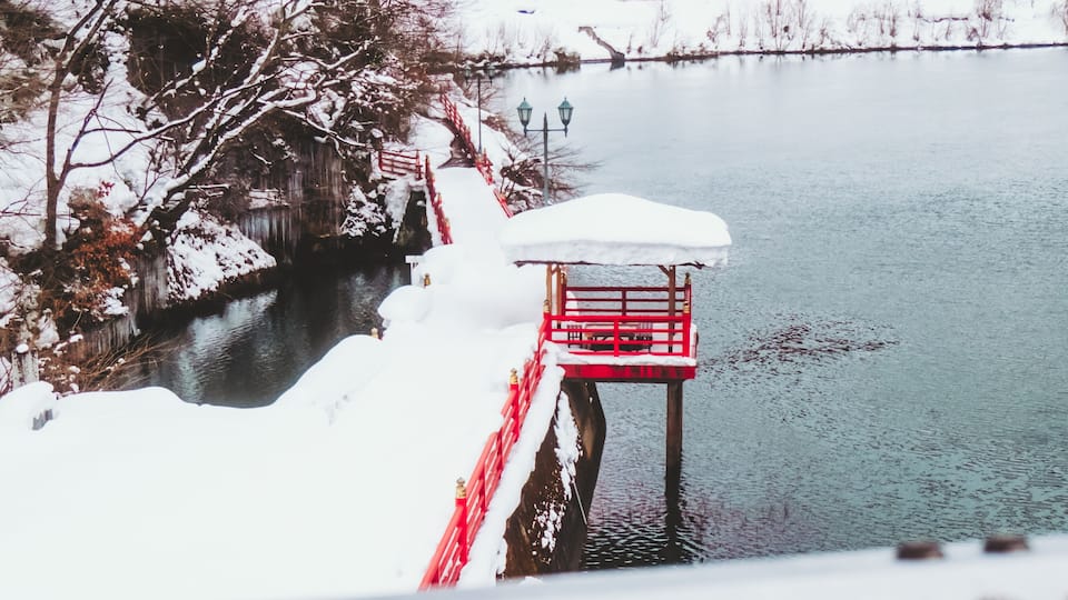 A snow-covered pier with a red structure, a bridge in the background, and a calm river.