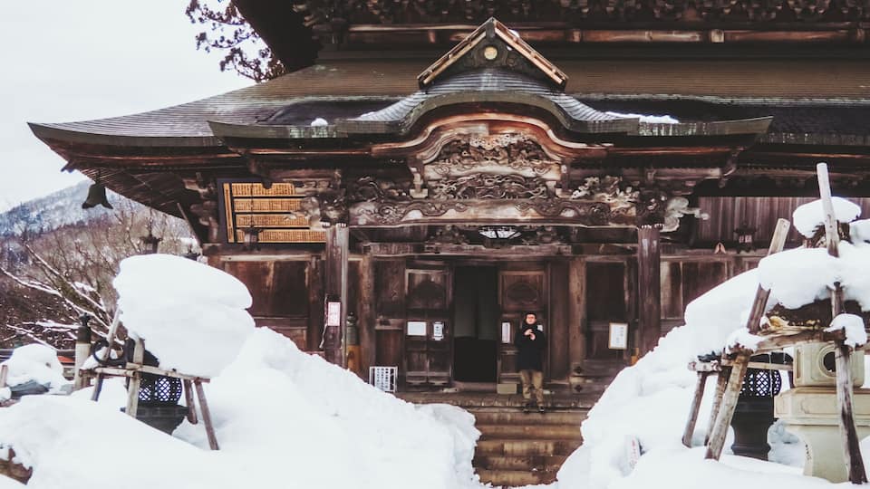 A traditional wooden building with intricate carvings, surrounded by snow-covered steps and trees.