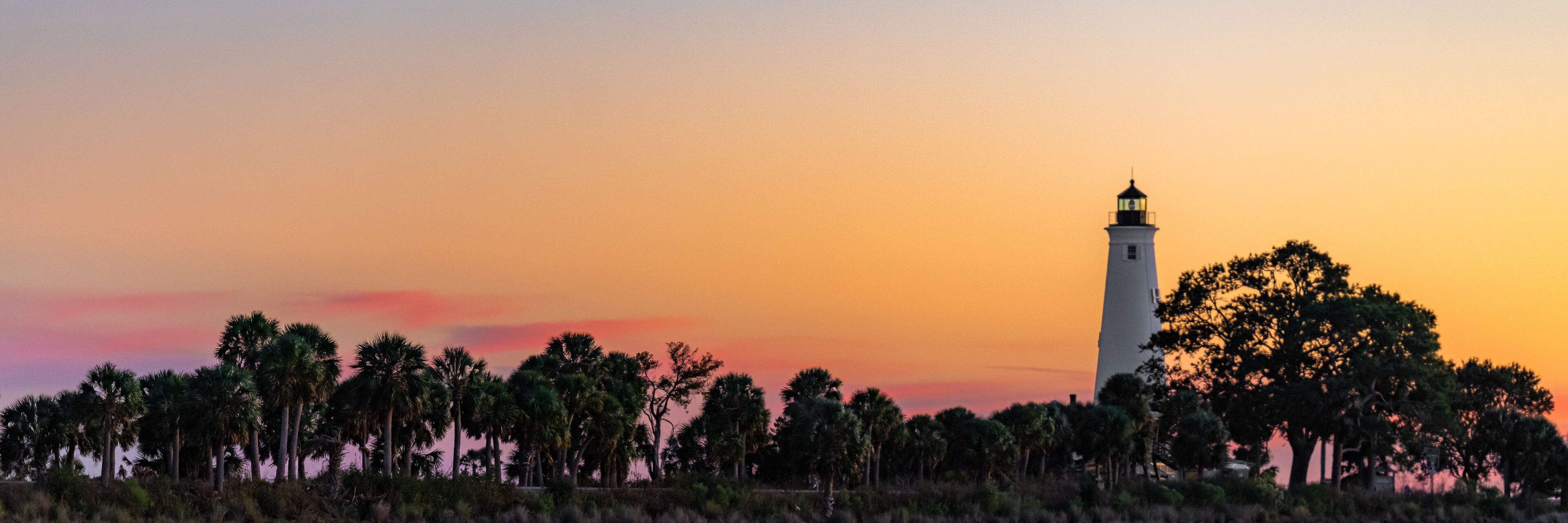 A lighthouse at sunset with palm trees in the foreground.