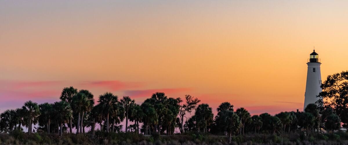 A lighthouse at sunset with palm trees in the foreground.