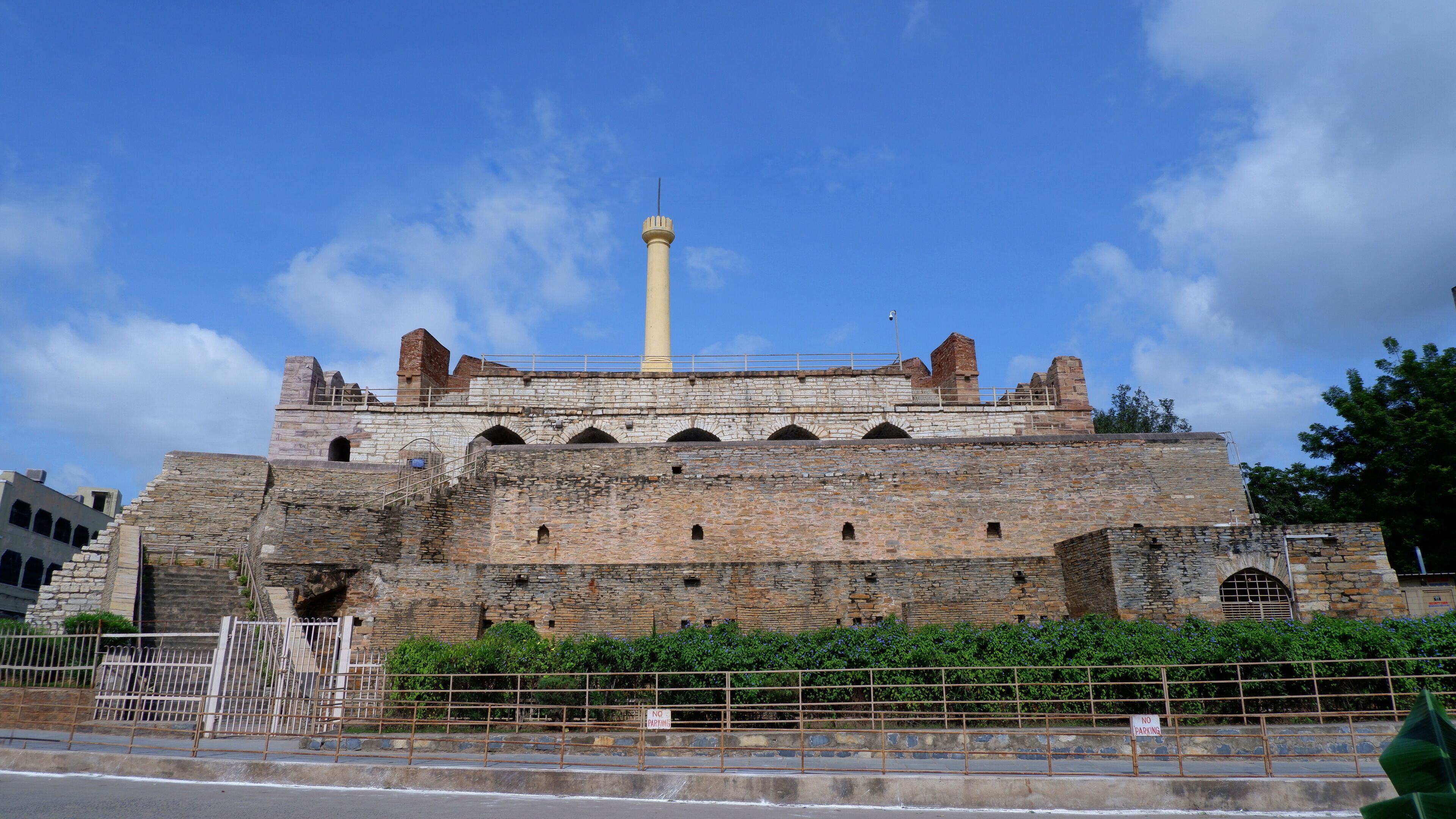 A historic stone fortress with a tall minaret, surrounded by greenery and a metal fence.