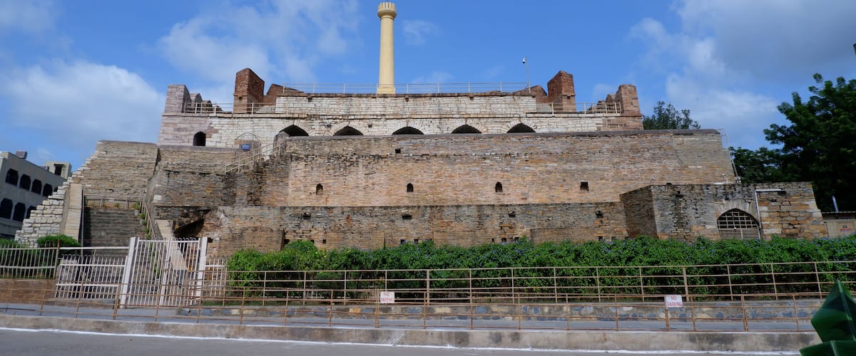 A historic stone fortress with a tall minaret, surrounded by greenery and a metal fence.