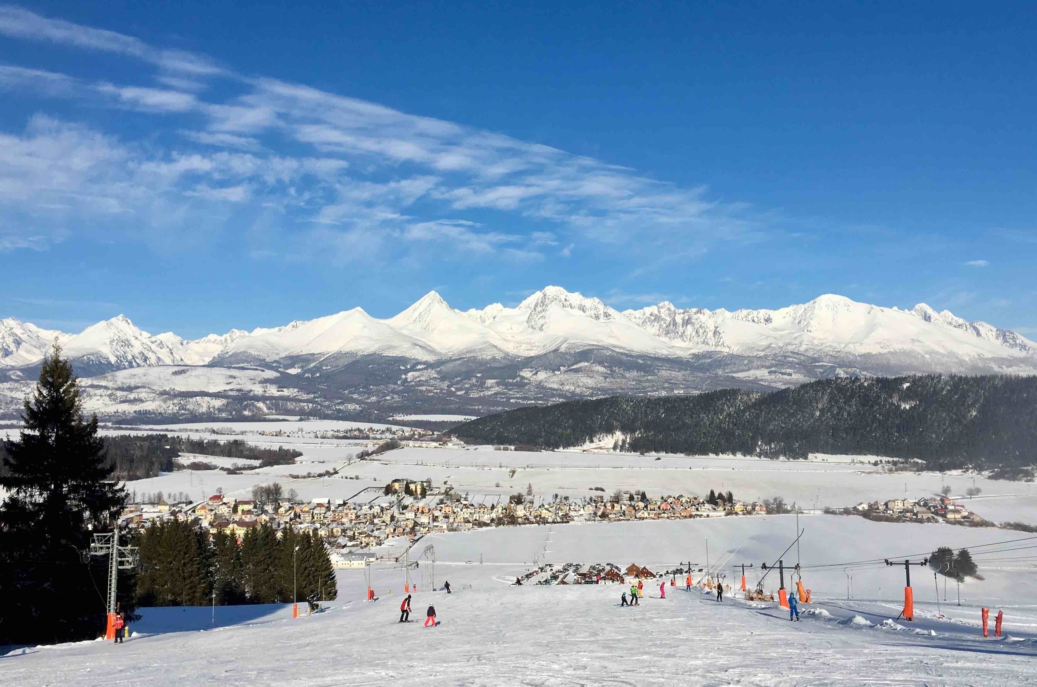 A snow-covered ski slope with a town in the valley and snow-capped mountains in the background.