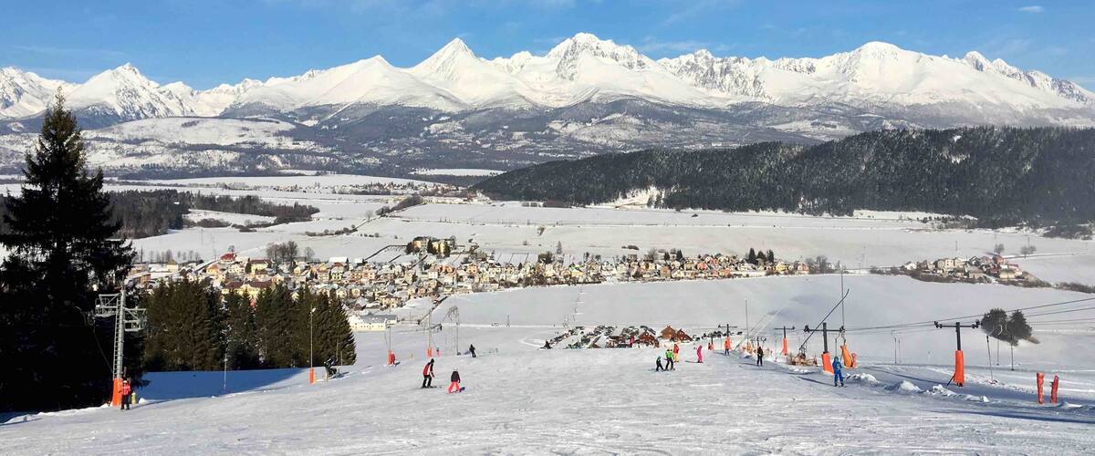 A snow-covered ski slope with a town in the valley and snow-capped mountains in the background.