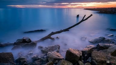 Um pôr do sol sobre um lago calmo, com um galho de árvore caído em primeiro plano e margem rochosa.