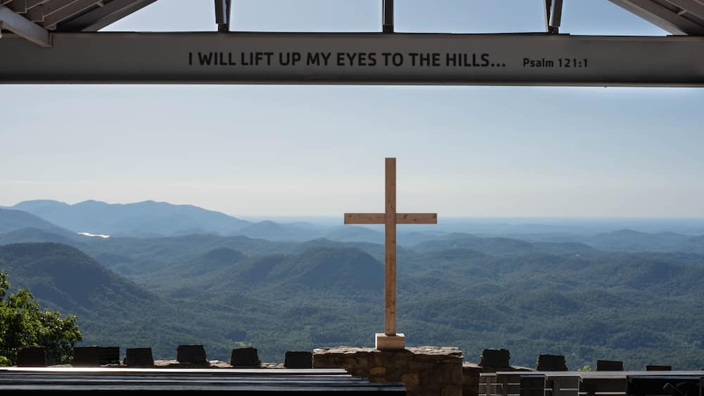 A wooden cross with a sign above it reading 'I WILL LIFT UP MY EYES TO THE HILLS... Psalm 121:1' against a backdrop of a mountainous landscape.