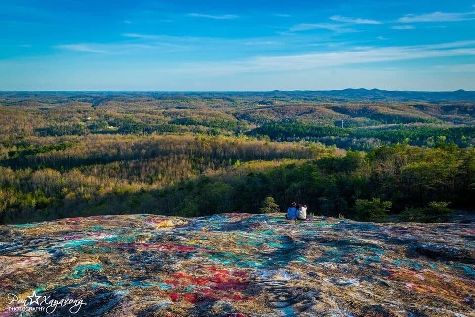 A vast landscape with a colorful rock formation in the foreground and a forested valley in the background.