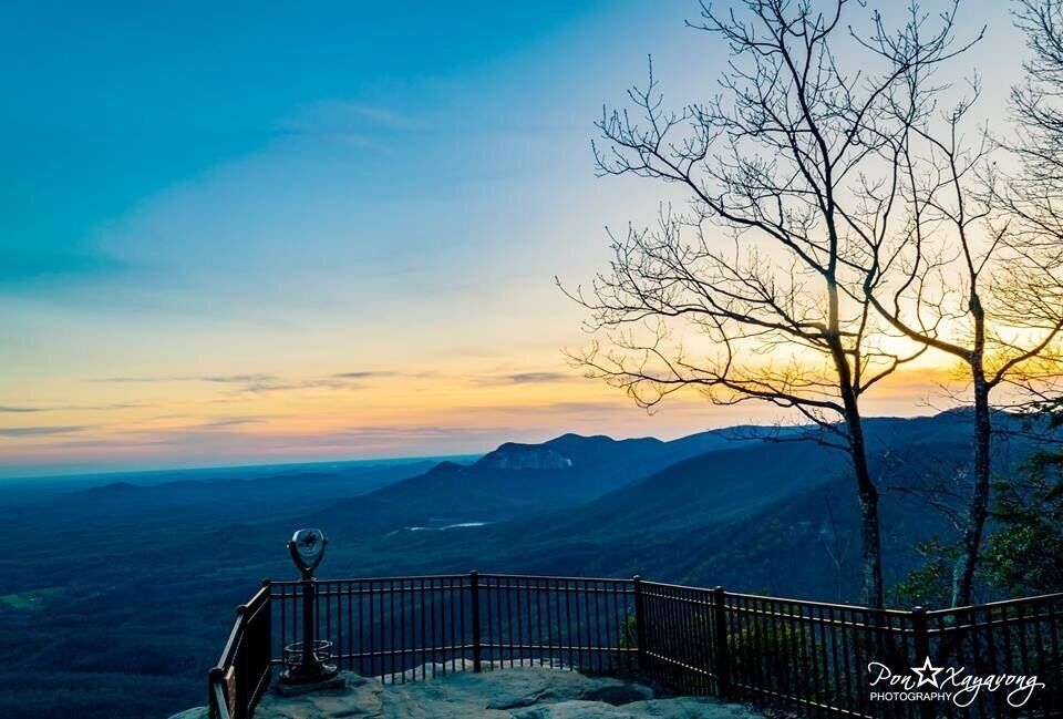A mountain landscape with a clear sky at sunset, a metal railing, and a leafless tree.
