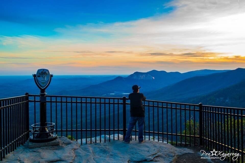 A person standing at a viewpoint with a telescope, overlooking a mountainous landscape during sunset.