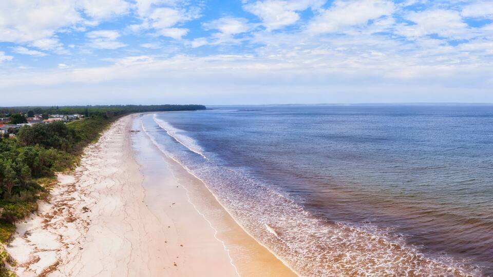 A coastal landscape with a sandy beach, gentle waves, and a distant shoreline with buildings.