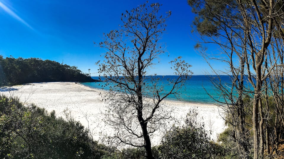 A beach with clear turquoise water, surrounded by trees and hills.