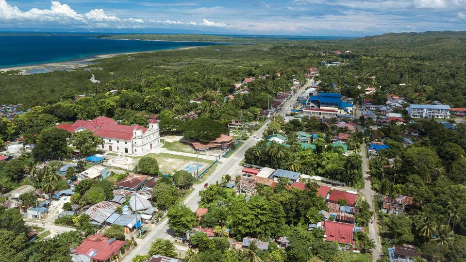 A coastal town with a prominent white building with a red roof, surrounded by lush greenery and a clear blue sky.