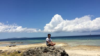 A person sitting on a rock by a beach with clear blue water and white clouds in the sky.