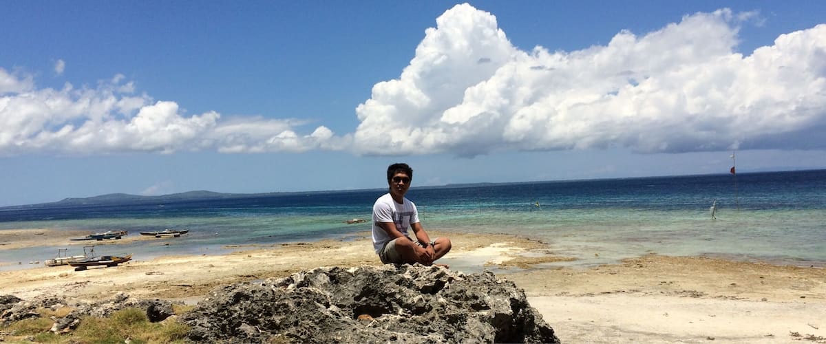 A person sitting on a rock by a beach with clear blue water and white clouds in the sky.