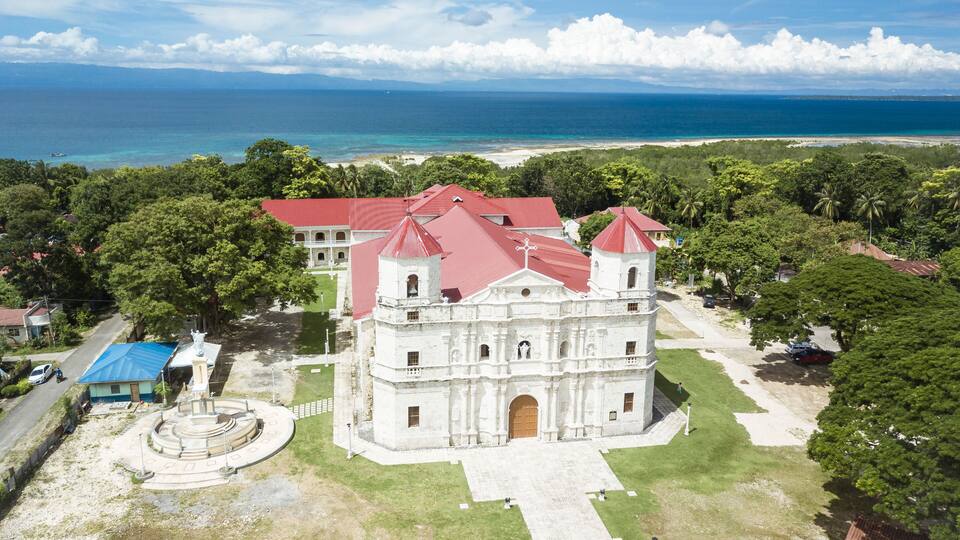 A white colonial-style building with red roofs, surrounded by lush greenery and a beach in the background.