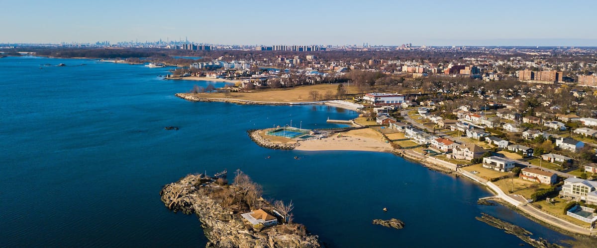 An aerial view of a coastal town with a river, a small island, and a distant city skyline.