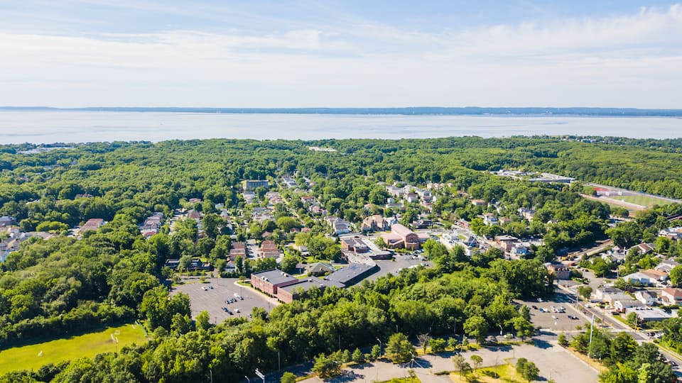 A town with dense green trees, a large body of water in the background, and a mix of residential and commercial buildings.