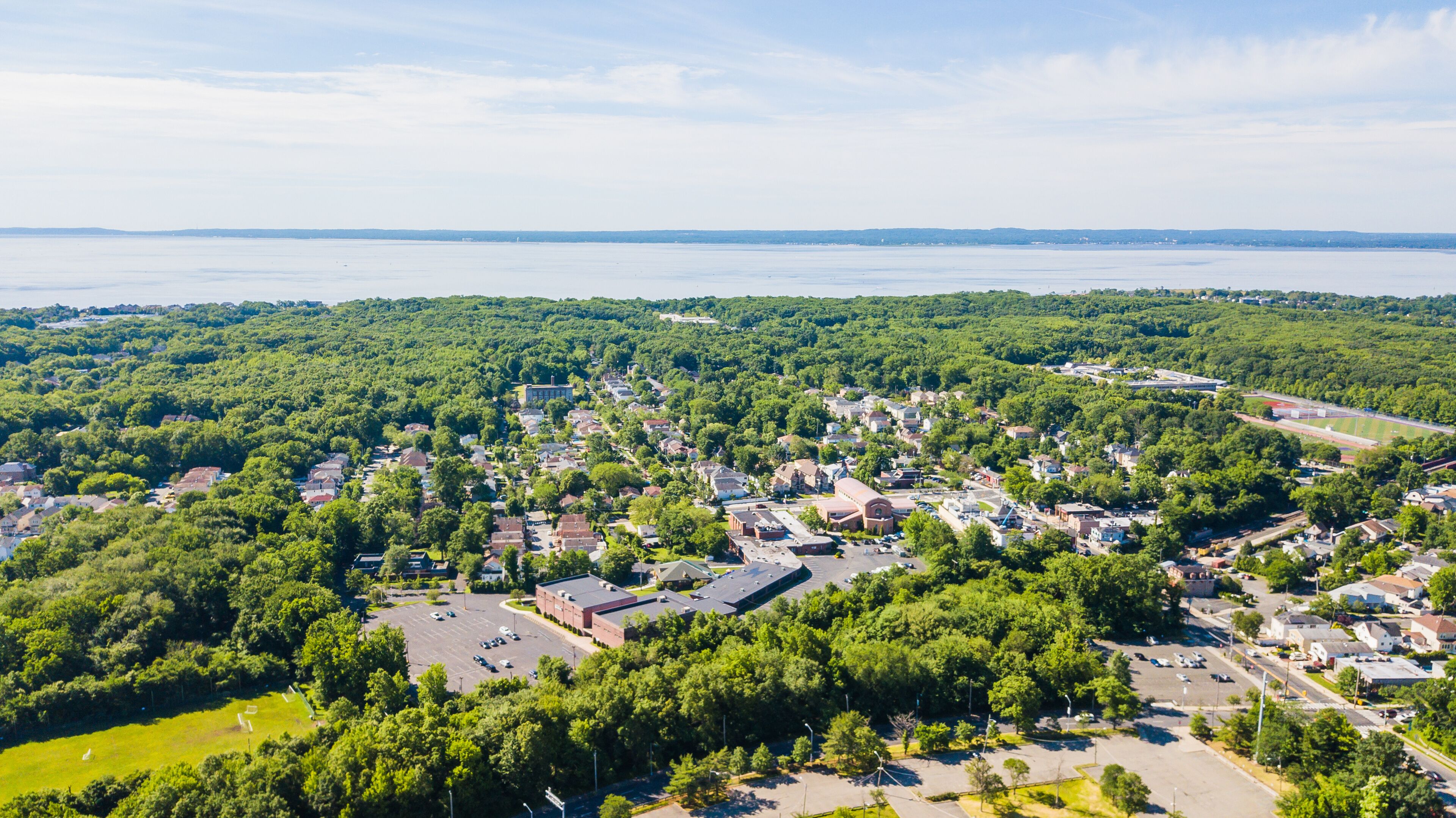A town with dense green trees, a large body of water in the background, and a mix of residential and commercial buildings.