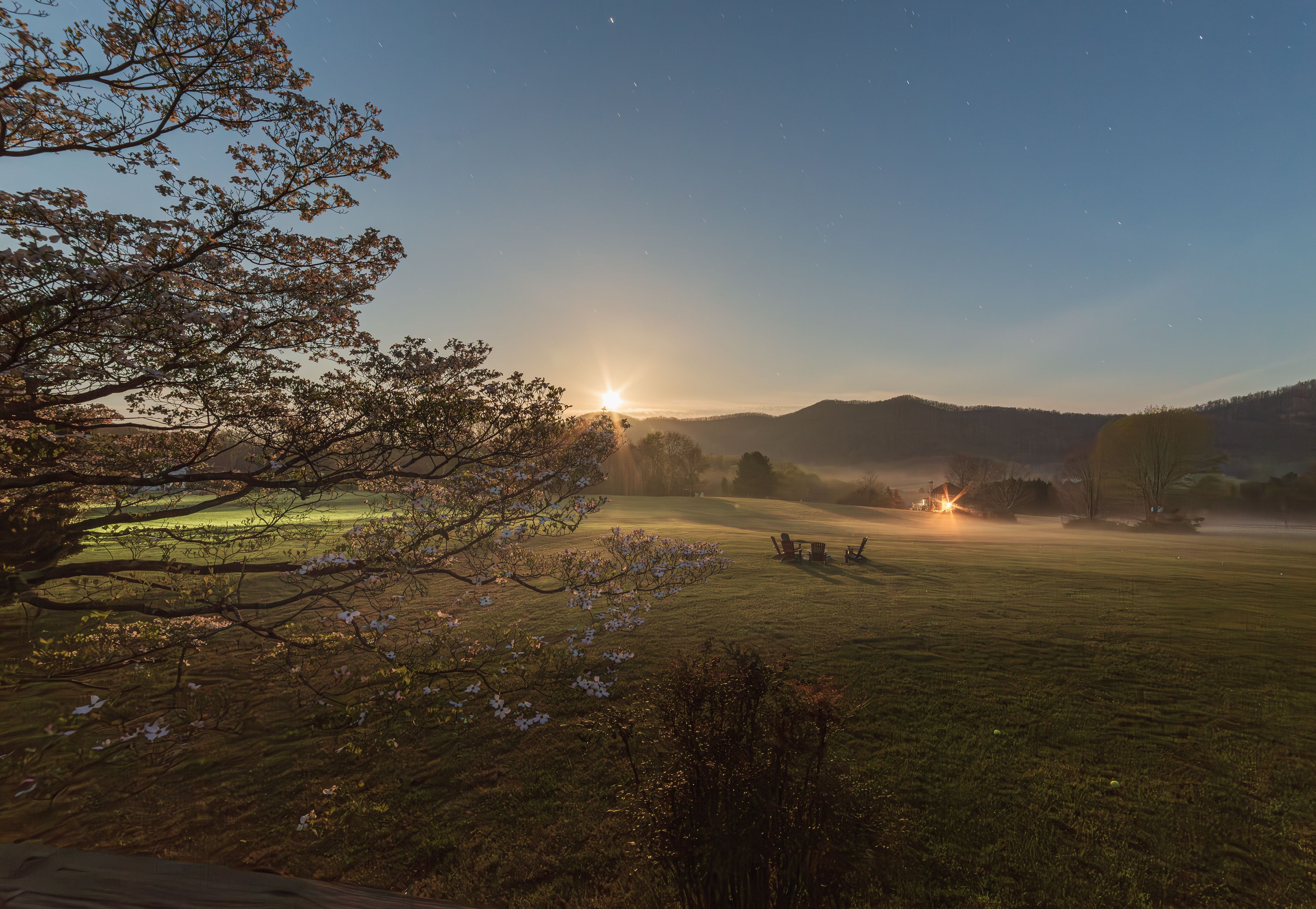 A sunrise over a grassy field with trees and a distant mountain range.