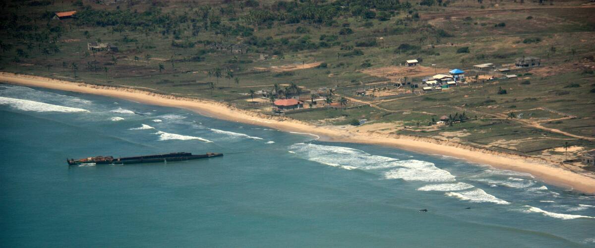A coastal landscape with a beach, ocean waves, a large ship, and scattered buildings.
