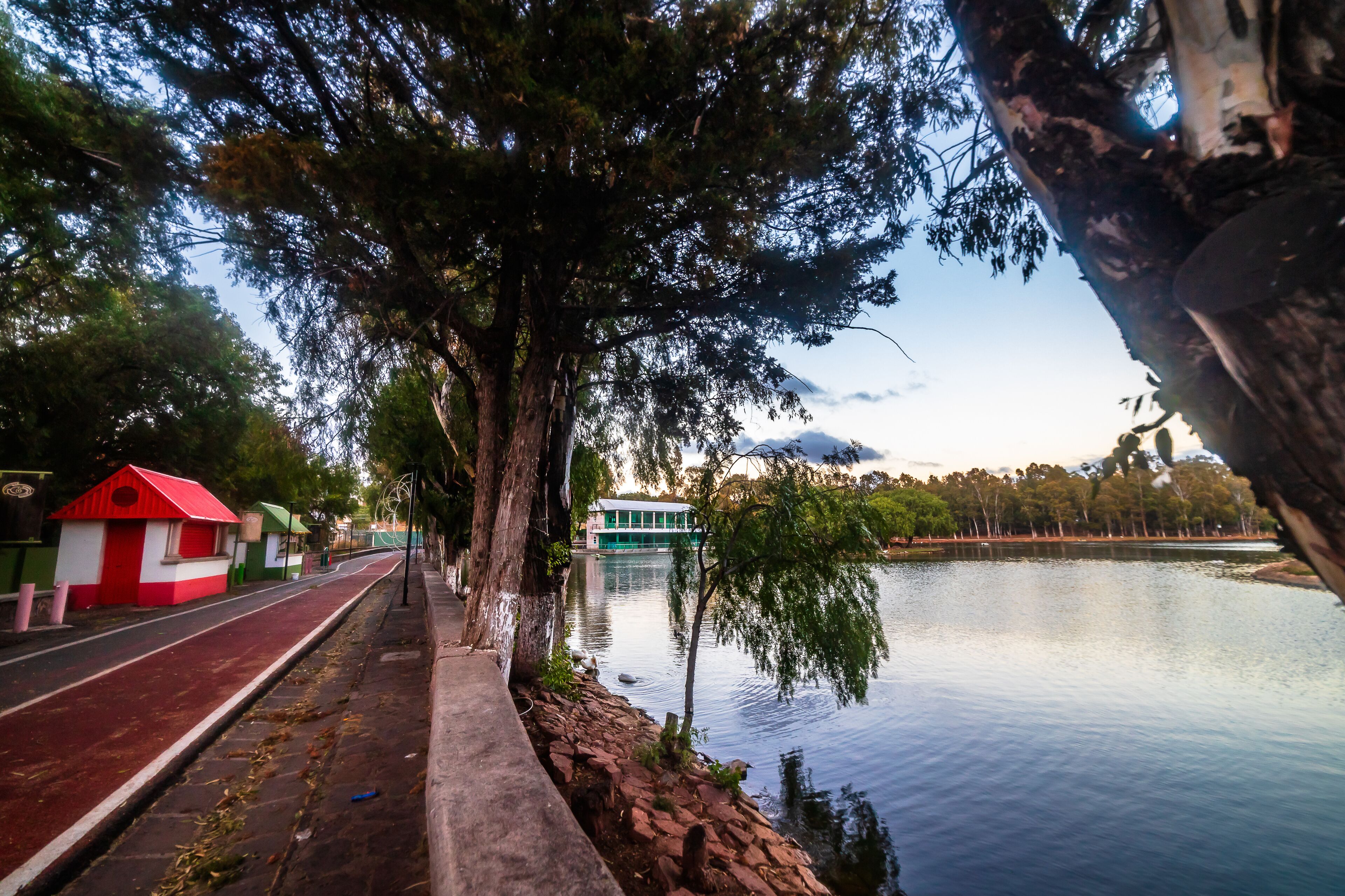 A riverside walkway with a red and white structure, a green building across the water, and trees lining the path.