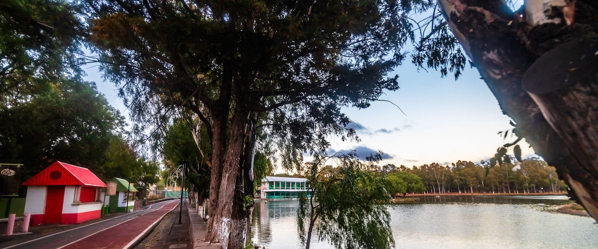 A riverside walkway with a red and white structure, a green building across the water, and trees lining the path.