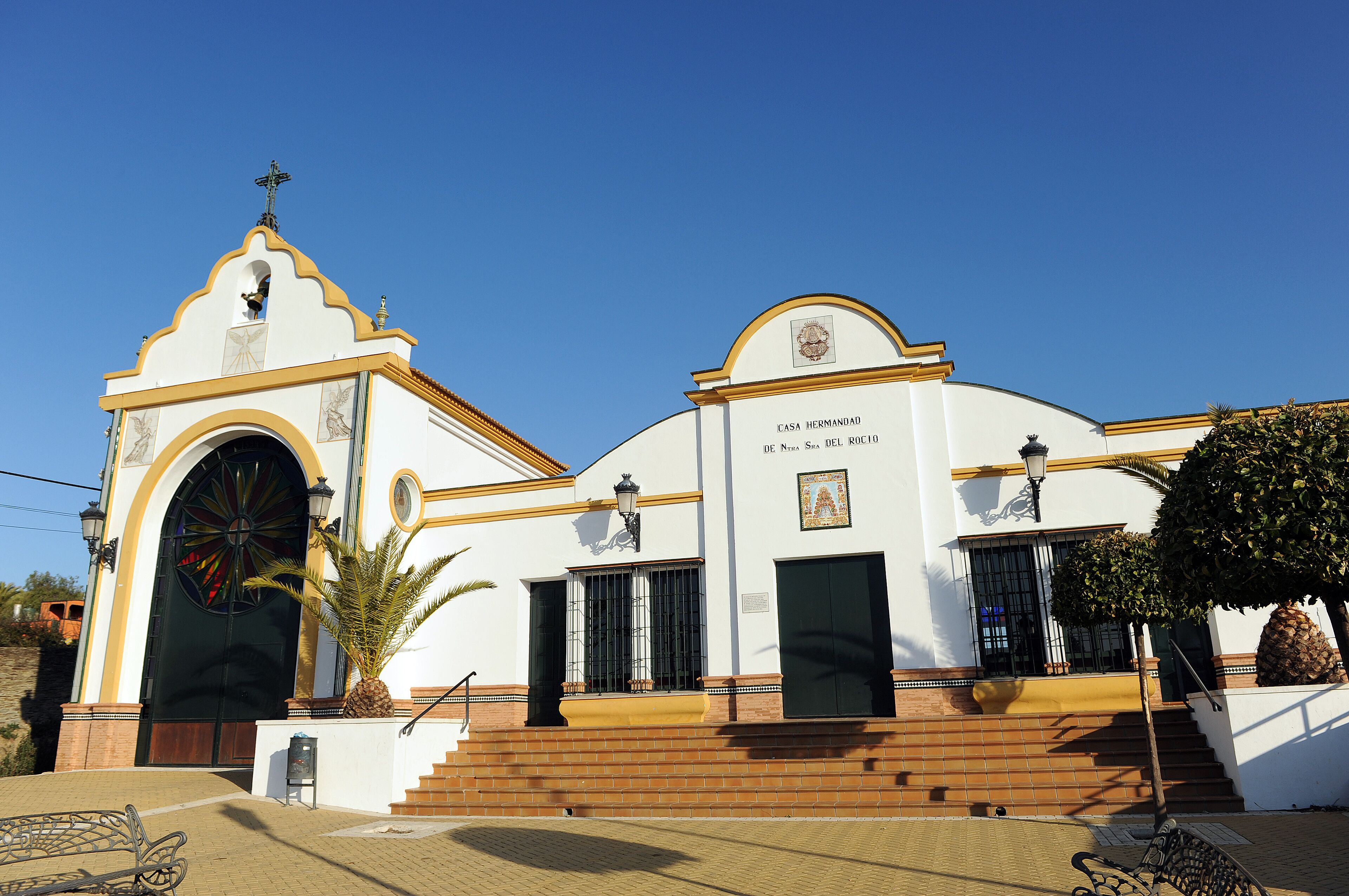 A white building with a cross on top, a large arched entrance, and a staircase leading up to the entrance.