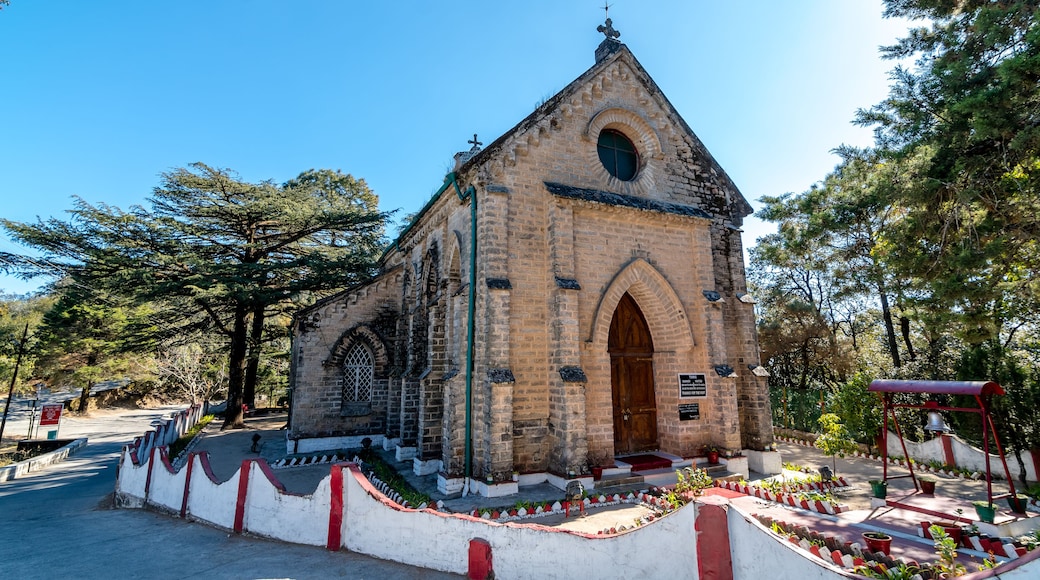 A small stone church with a bell tower, surrounded by trees and a white wall with red trim.